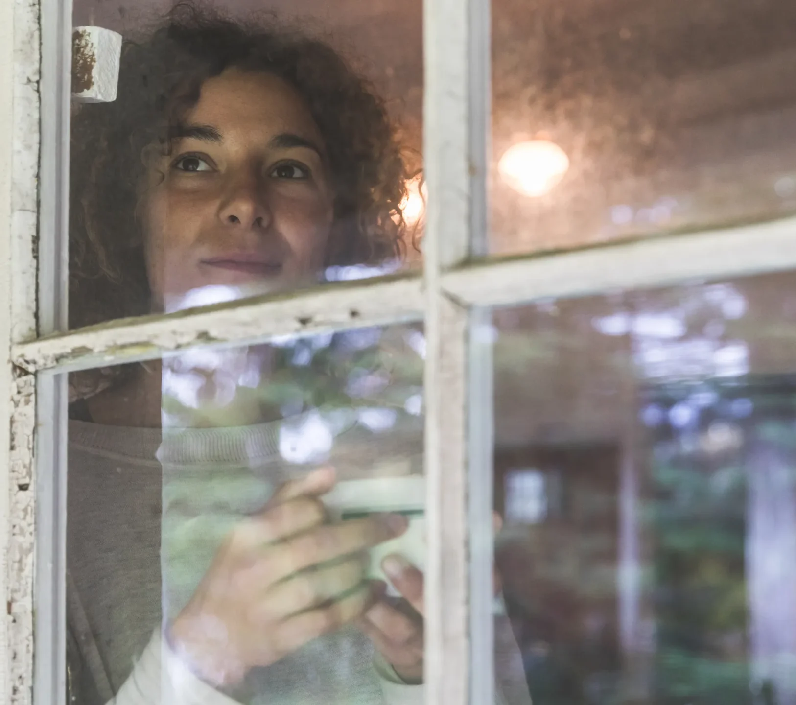 Women standing inside holding a cup of coffee and looking out the window
